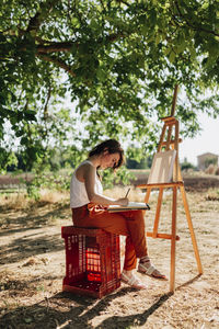 Young woman artist drawing in spiral notebook while sitting next to easel