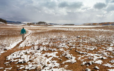 Rear view of man walking on snow covered field against cloudy sky