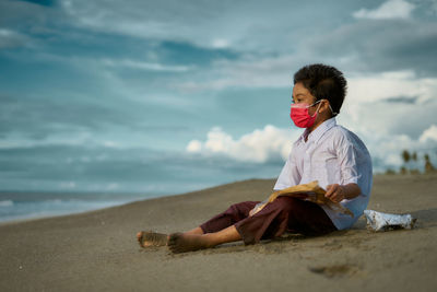 Boy wearing mask sitting at beach against sky