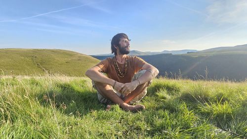 Young man sitting on grassy field against sky