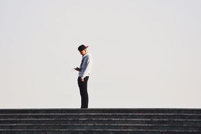 Young man using on mobile phone while standing on steps against clear sky