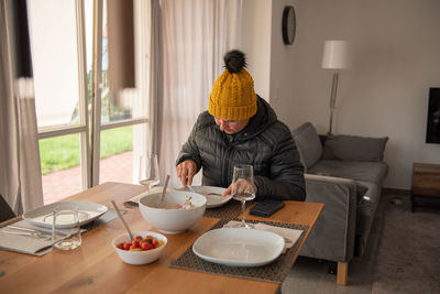 A woman sits at the dining table, dressed warmly, and eats