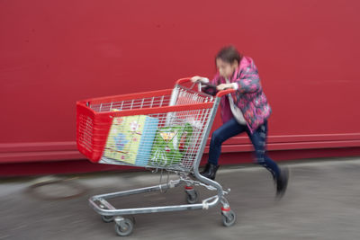 Playful girl pushing shopping cart against red wall