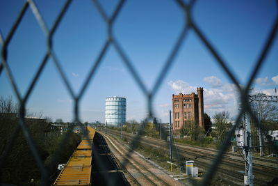 Railroad tracks in city against sky seen through chainlink fence