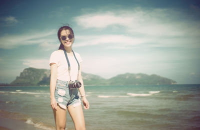 Portrait of smiling young woman with camera standing at shore of beach 