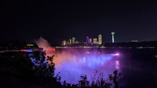 Illuminated buildings by lake against sky at night