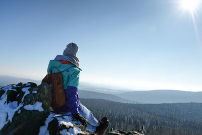 Rear view of man sitting on mountain against sky during winter