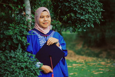 Smiling young woman wearing graduation gown in forest
