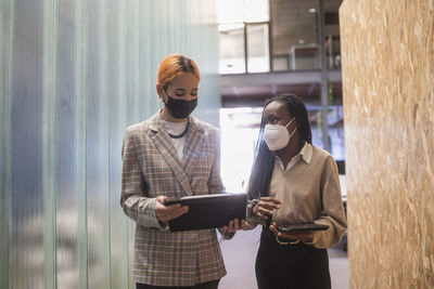 Multiethnic female colleagues with tablets and in protective masks standing in coworking space and discussing project