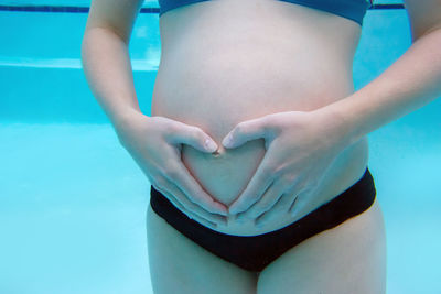 Midsection of pregnant woman making heart shape with hands on stomach while swimming in pool