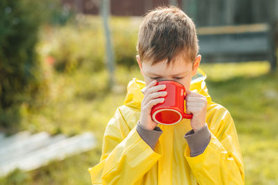 A little boy in a yellow raincoat is drinking from a red mug on the street. camping concept. 