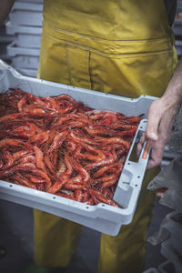 Crop man in uniform carrying box with fresh red shrimps in market stall