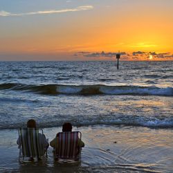 Rear view of friends sitting on deck chairs at beach against sky during sunset