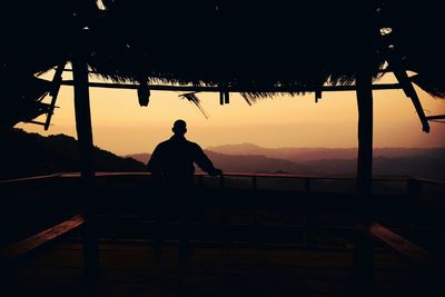 Rear view of silhouette man standing by railing against sky during sunset