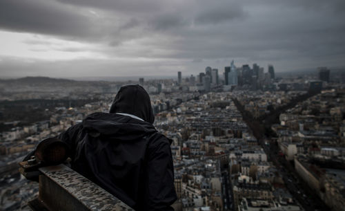 Rear view of man looking at cityscape while sitting on building terrace against sky