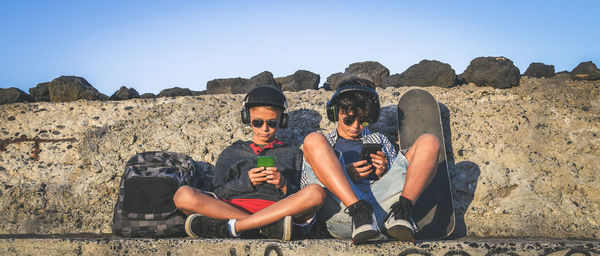 Friends listening music on mobile phone while sitting on rock against clear sky