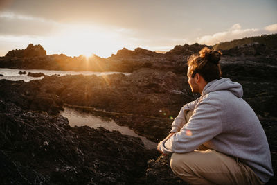 Rear view of woman sitting on rock against sky during sunset