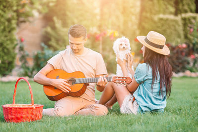 Man playing guitar while woman sitting with dog at park