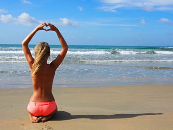 Rear view of shirtless woman making heart shape while kneeling on shore at beach