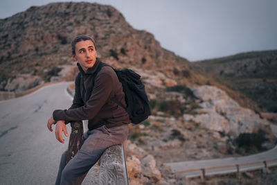 Young man exploring mountains while sitting on railing at sunset