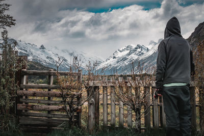 Rear view of man standing by fence against snowcapped mountains