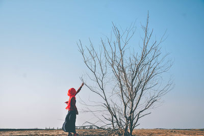 Side view of young woman touching bare tree branch against clear sky