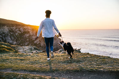 Back view of unrecognizable african american female owner running with border collie dog while spending time together on beach near waving sea at sunset