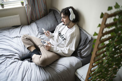 Man using mobile phone while holding game controller in bedroom