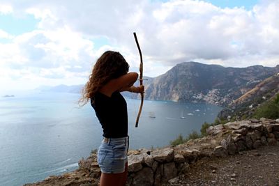 Side view of woman practicing archery on cliff against sea