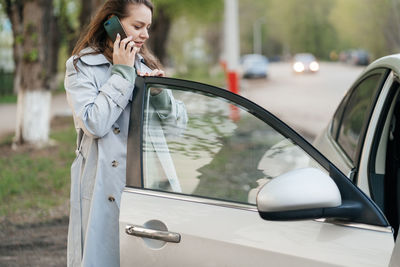 Beautiful girl with long hair in a grey trench coat using smartphone call gets into the car