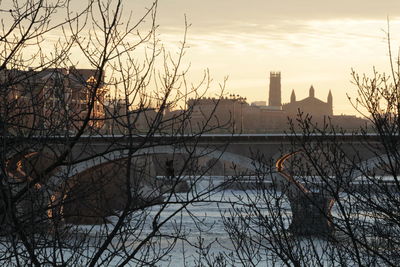 Bare trees and buildings against sky during sunset