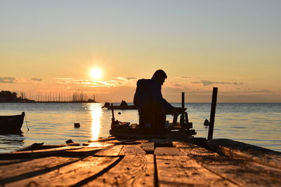 Silhouette man sitting by sea against sky during sunset