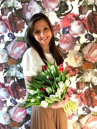 Portrait of smiling young woman holding flowers while standing against wall