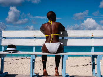 Rear view of woman in bikini sitting on beach against sky