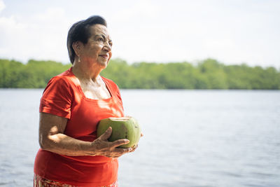 Old hispanic or latina woman holding a coconut next to a lake, river or estuary with mangroves.