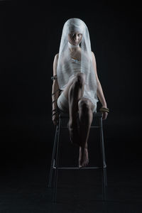 Full length of woman wrapped in bandage sitting on chair against black background