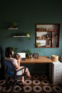 Side view of asian female freelancer reading documents on laptop while sitting and drinking tea at table during remote work