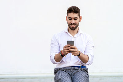 Young bearded man sitting against a white wall using the mobile phone