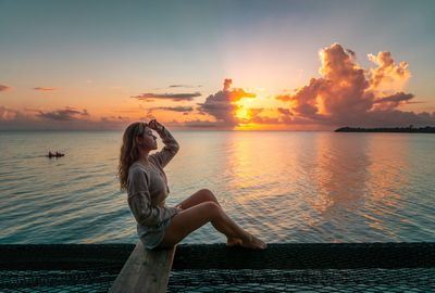 Side view of woman sitting at beach against sky during sunrise