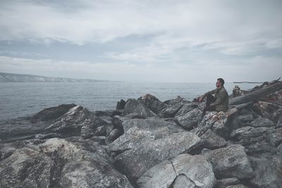 Side view of man looking at sea while sitting on rocks against cloudy sky