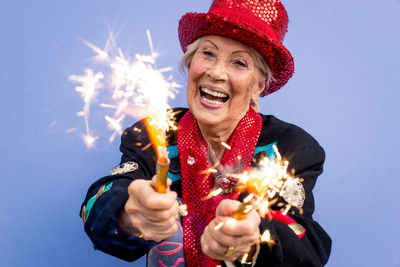 Portrait of happy senior woman wearing hat holding sparklers against blue background