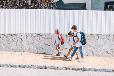 Side view of schoolboy with backpack speaking with female friends while strolling on tiled pavement against stone wall in sunlight