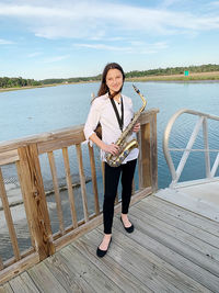 Portrait of young woman holding a saxophone standing by railing against the ocean  