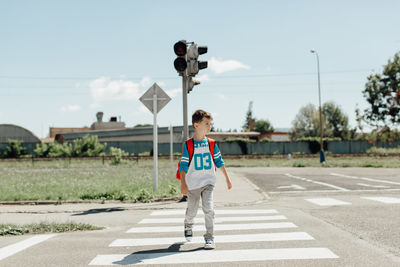 Full length of boy with backpack crossing road against sky during sunny day