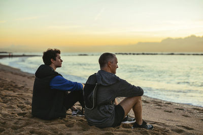 Thoughtful father and son resting while sitting t beach against sky during sunset