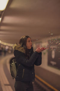Young woman blowing confetti while standing against train at subway station