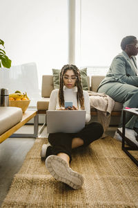 Female programmer using smart phone while sitting with laptop on carpet in office