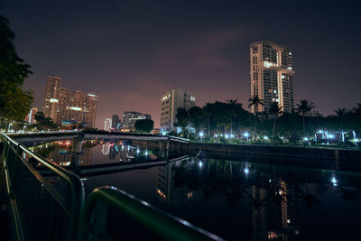 Illuminated modern buildings by river against sky at night