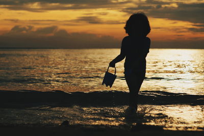 Silhouette boy standing on beach against sky during sunset