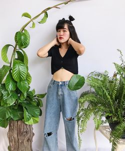 Portrait of woman wearing black crop top and jeans standing beside plant